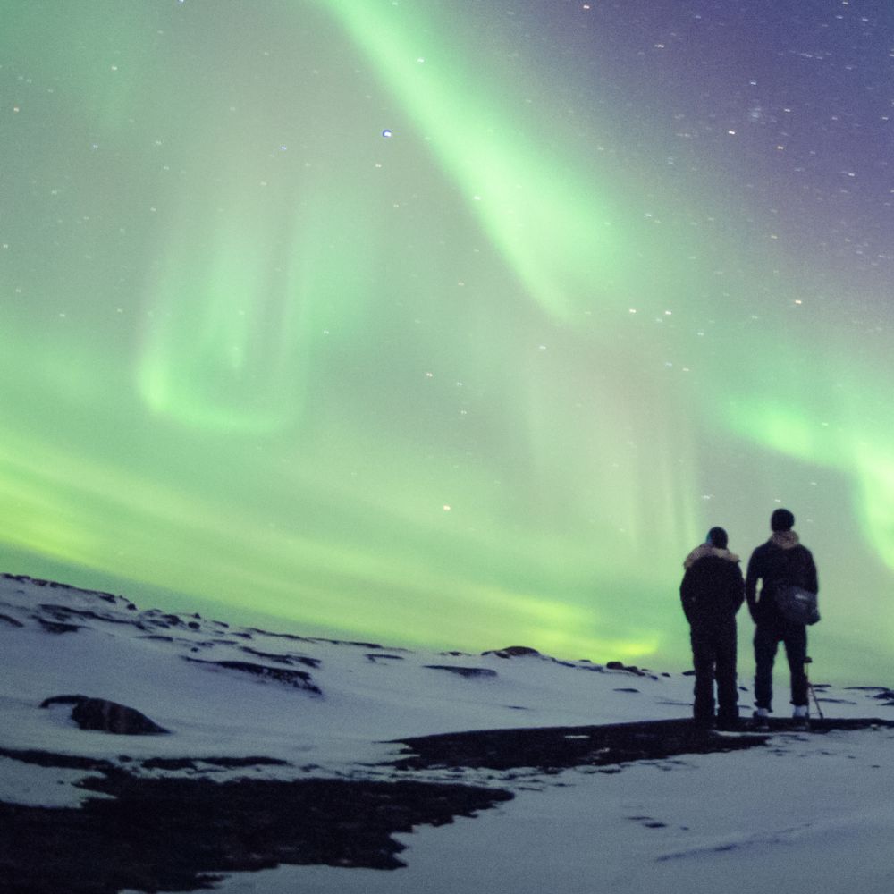 Silhouette of two people standing on a snowy ridge looking at the green and purple Northern Lights.