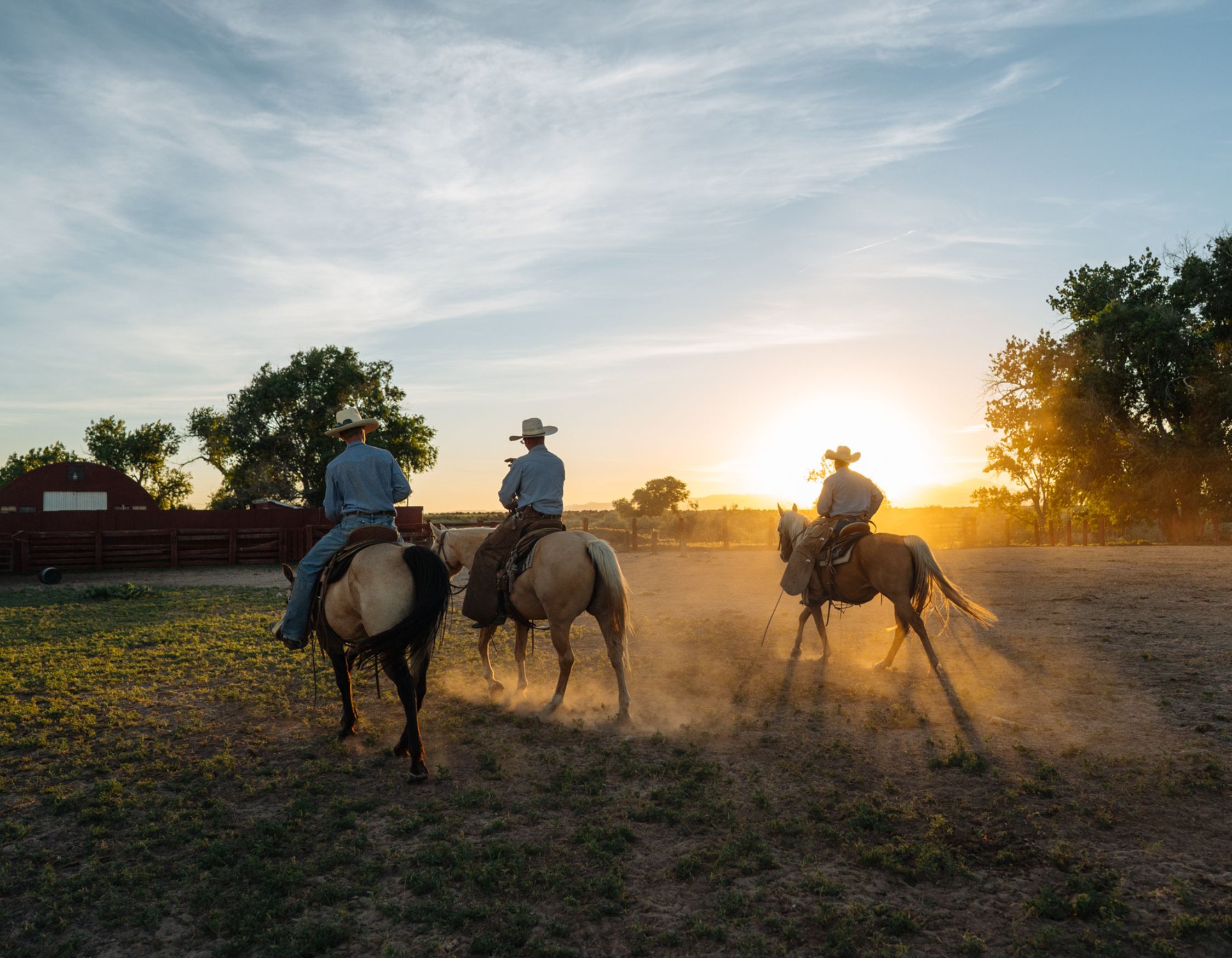 Three cowboys on horseback riding away at sunset, kicking up dust in an open field. A red barn is visible in the background, surrounded by trees.