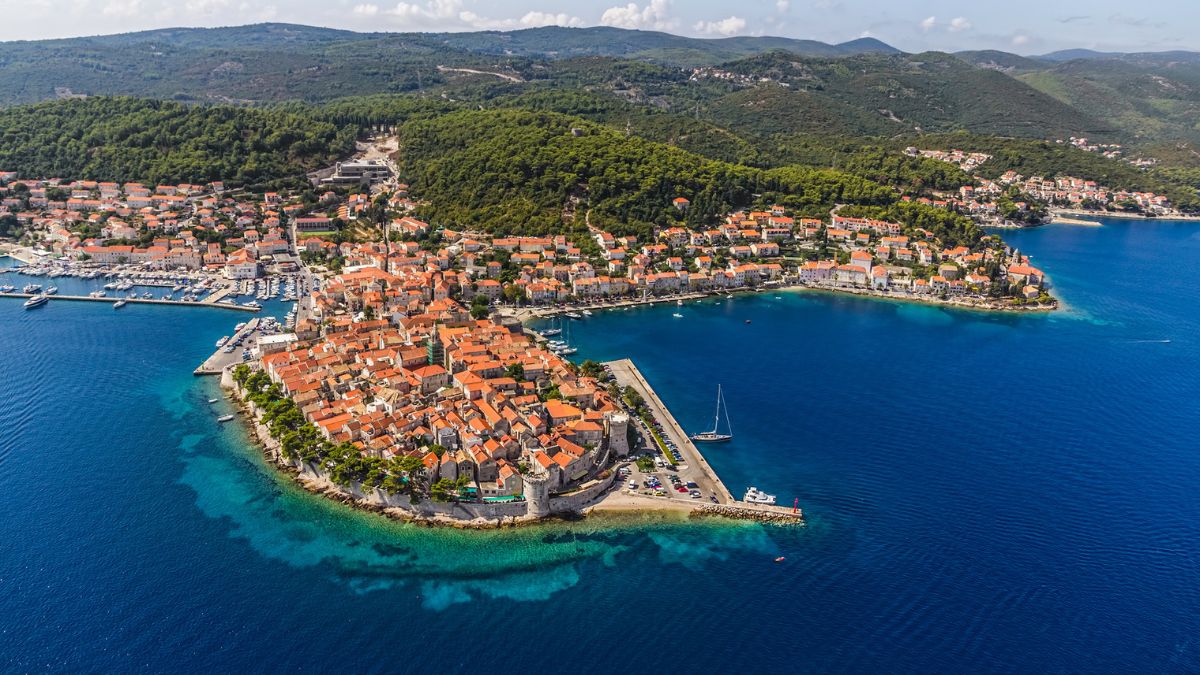 Aerial view of a coastal town surrounded by clear blue waters, featuring red-tiled roofs and lush green hills in the background. The shoreline includes a marina with several boats.