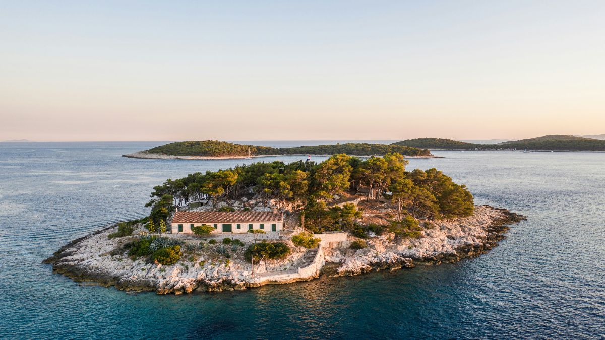 Aerial view of a small rocky island with a single long white building and green trees surrounded by blue ocean.
