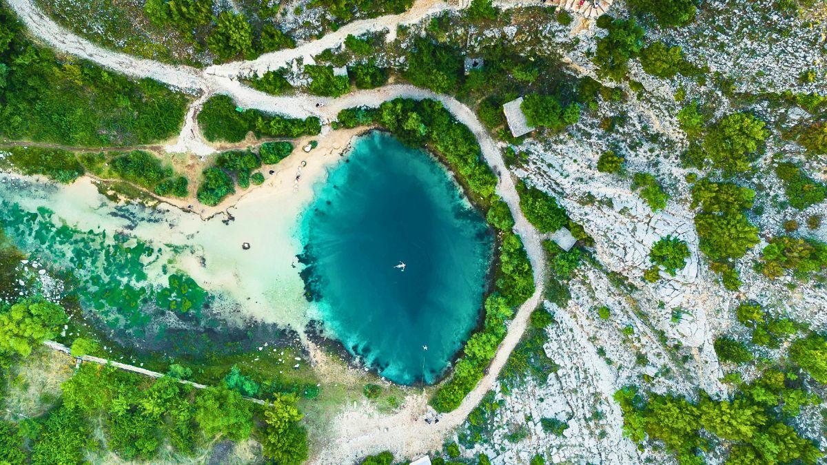 Aerial view of a round, vibrant blue lagoon surrounded by lush greenery and rocky terrain. The water appears clear and inviting, with a few small rocks and a path visible nearby.