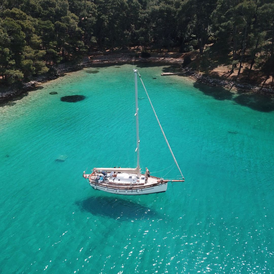 A small sailing boat anchored in clear turquoise waters, surrounded by lush greenery and rocky shores.