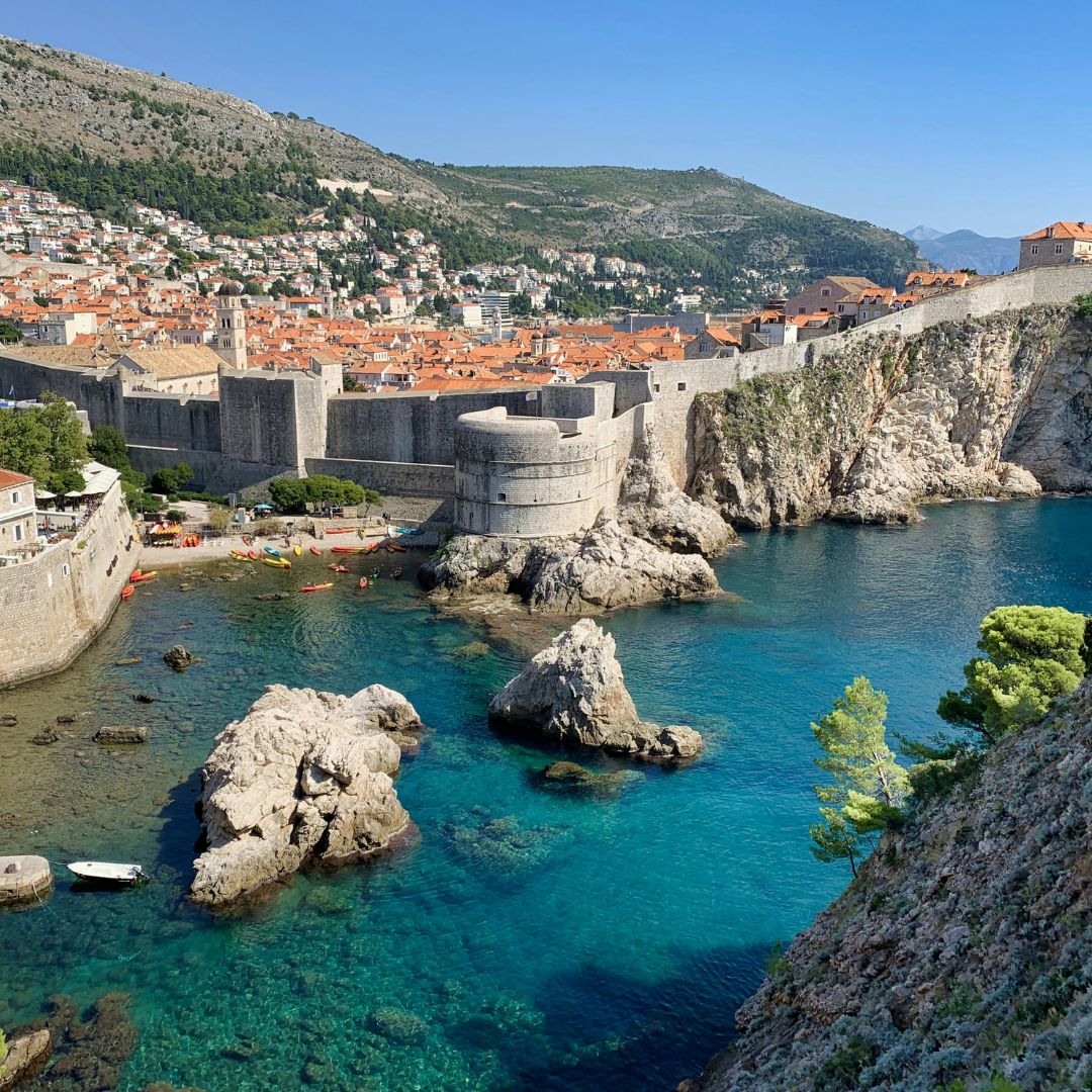 A scenic view of Dubrovnik's city walls and a historic tower, surrounded by vibrant blue waters. Rocky formations and small boats are visible in the foreground, while the terracotta rooftops of the city are nestled against the hillside in the background. Lush greenery adds to the natural beauty of the landscape.