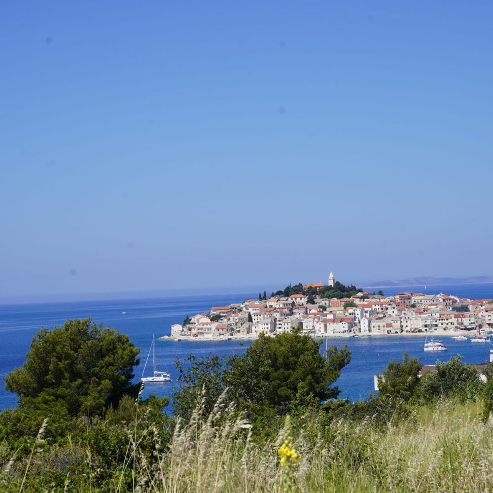 A coastal view of a picturesque island town with colorful buildings and a lighthouse, surrounded by clear blue water and boats. Lush greenery is visible in the foreground, under a bright blue sky.