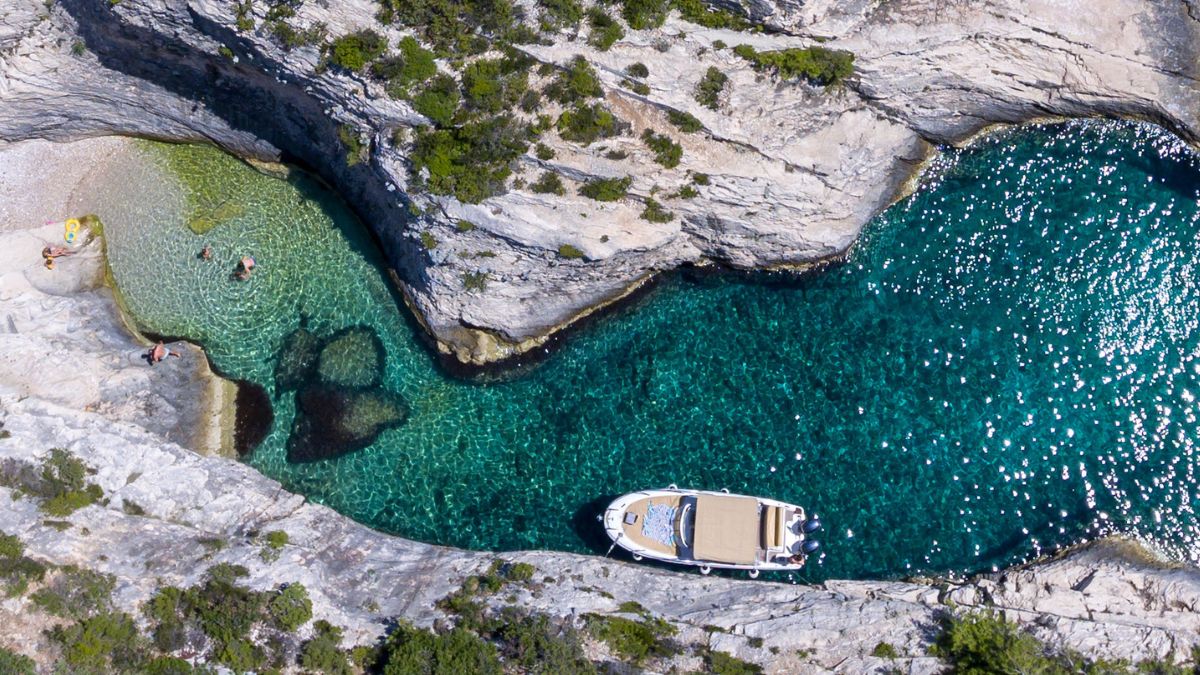 Aerial view of a serene coastline featuring a small boat anchored in crystal-clear waters. A secluded cove with sandy beach and people enjoying the sun is visible nearby, surrounded by rocky cliffs and lush greenery.