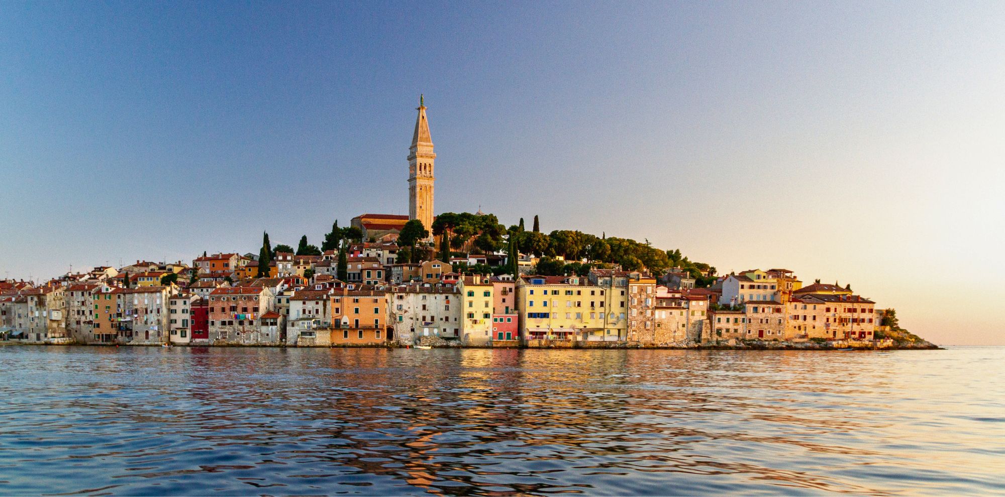 A picturesque coastal town with colorful buildings lining the waterfront, leading up to a tall clock tower on a hill. The scene is illuminated by the warm glow of sunset, reflecting in the calm water below.