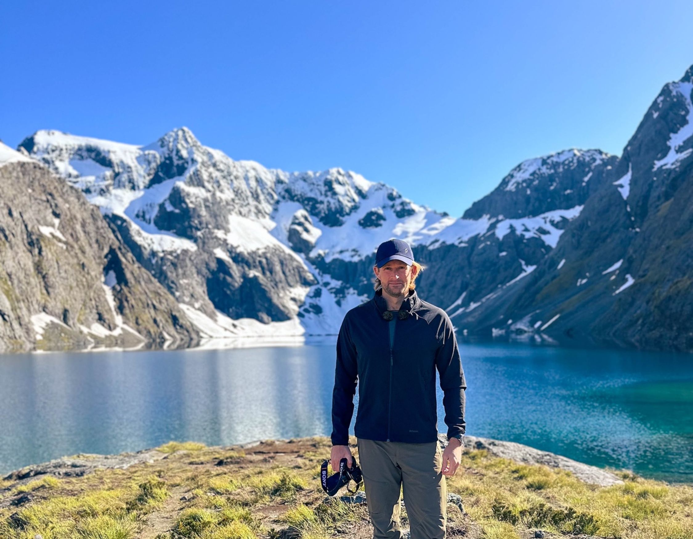 Pelorus founder, Jimmy Carroll, in hiking gear and a blue cap standing on a grassy ridge overlooking a clear mountain lake.