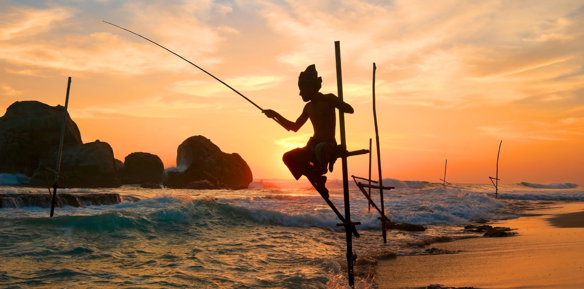 A silhouette of a fisherman perched on a wooden stilt, casting a line into the ocean at sunset. The vibrant colors of the sky reflect orange and pink hues as waves gently crash against the shore. Large rocks are visible in the background, creating a tranquil and picturesque scene.