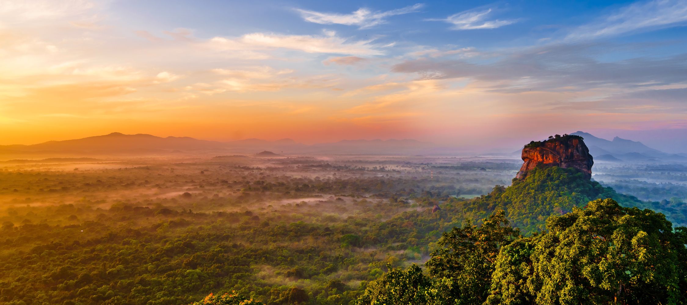 A panoramic view of a misty landscape at sunrise, featuring a prominent rocky hill surrounded by lush greenery and rolling hills in the distance. The sky is painted with warm hues of orange and pink, creating a serene atmosphere.
