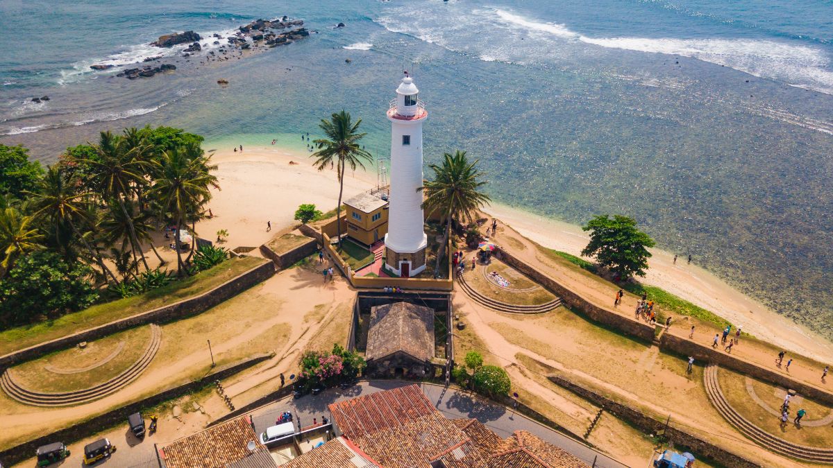 Aerial view of a coastal lighthouse surrounded by palm trees, sandy beach, and clear blue water. People can be seen walking along the beach and near the lighthouse, with rocky formations visible offshore.