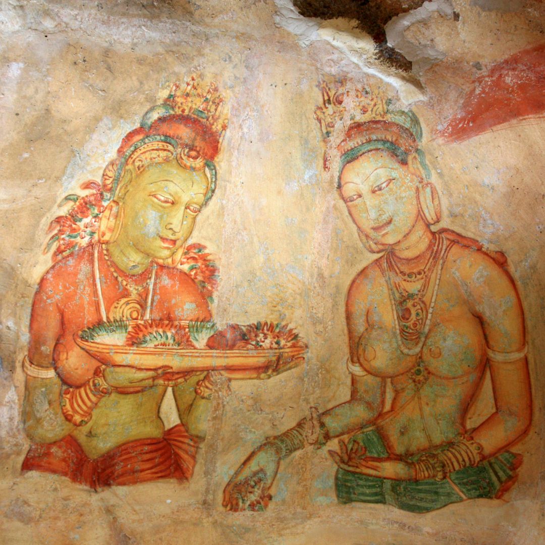 Two ancient Buddhist figures depicted in vibrant hues on a cave wall. The figure on the left holds a platter adorned with fruit, while the figure on the right gazes at her. Both are adorned with elaborate jewelry and hairstyles, showcasing intricate details.