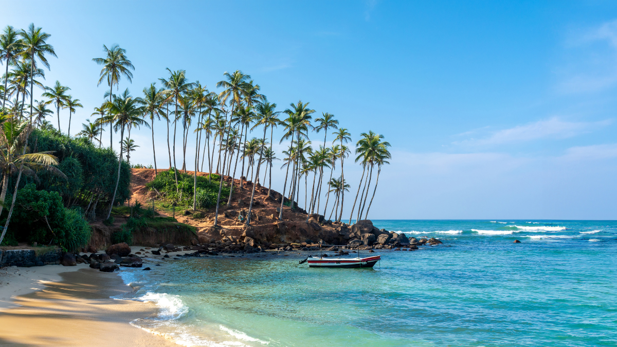 A serene beach scene with golden sand and clear turquoise water, bordered by tall palm trees. A small boat floats gently on the waves, with a rocky shoreline in the background under a bright blue sky.