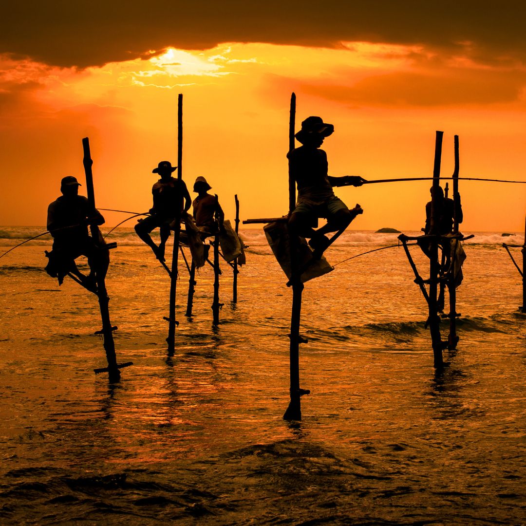 Silhouettes of fishermen perched on tall poles in the ocean during a dramatic sunset, casting shadows against a vibrant orange sky.