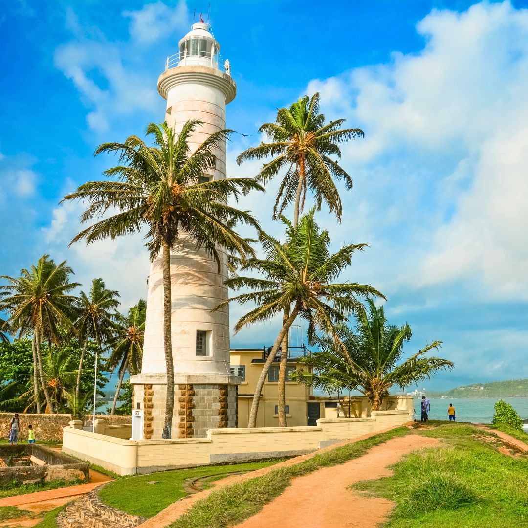 A tall white lighthouse surrounded by green palm trees, with a blue sky and fluffy clouds in the background. People walk along a sandy path nearby, with the ocean visible in the distance.