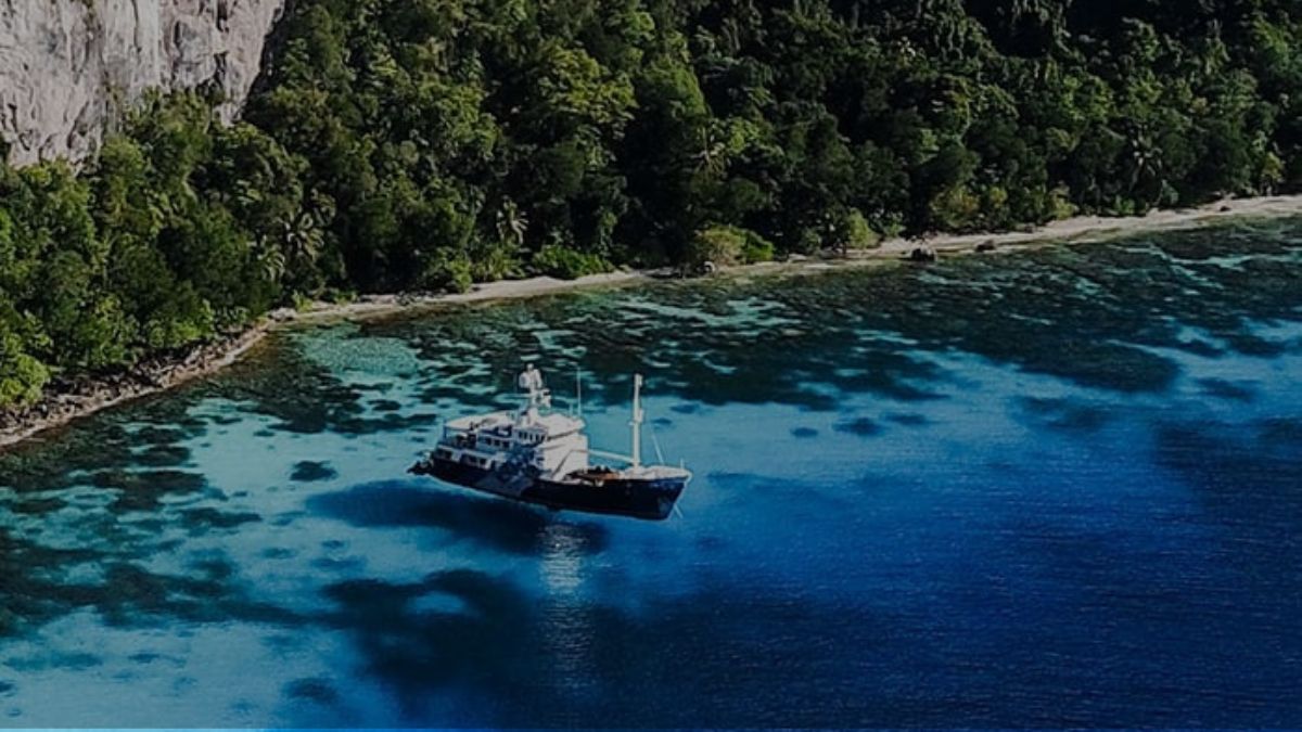 A research vessel anchored near a vibrant coral reef along a lush, tropical coastline. Clear blue waters reflect the surrounding greenery and rocky cliffs.