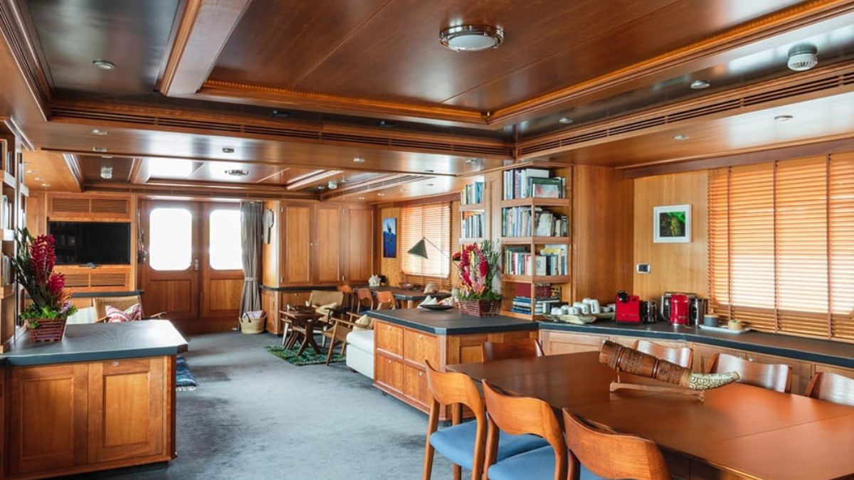A spacious, luxurious interior of a yacht featuring warm wooden finishes, a dining area with a large table and blue chairs, a cozy seating area with armchairs, and a well-equipped kitchen area. Sunlight filters through large windows, highlighting decorative elements and plants.