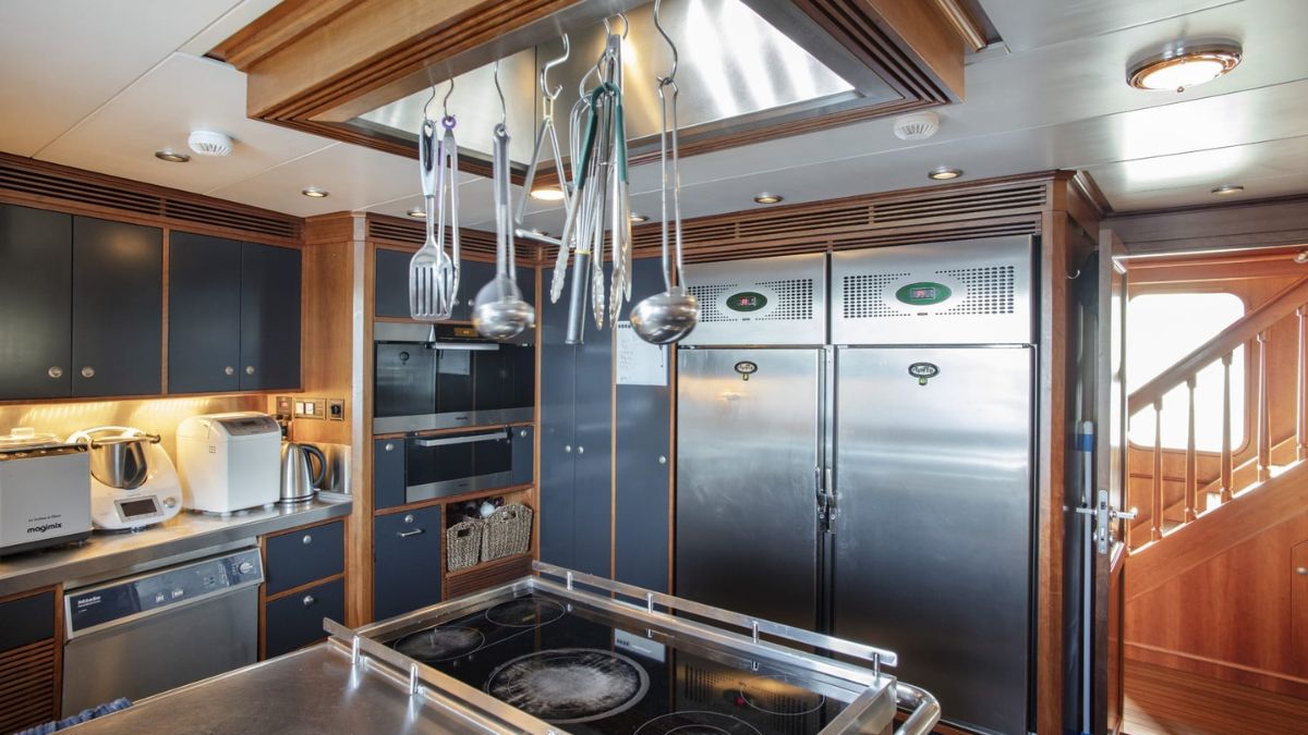 A modern kitchen featuring stainless steel appliances, including refrigerators and an oven. Cooking utensils hang from a rack above a central cooking island with stovetops. The cabinets are dark wood, and there is an elegant staircase in the background.