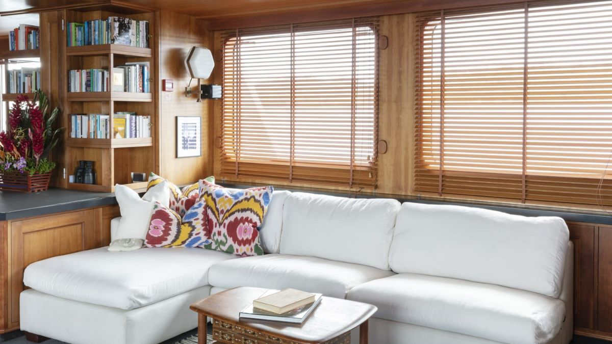 A cozy living area featuring a white sectional sofa adorned with colorful patterned pillows, a wooden coffee table with a book on it, and a bookshelf filled with books against a wooden wall with large windows covered by wooden blinds.