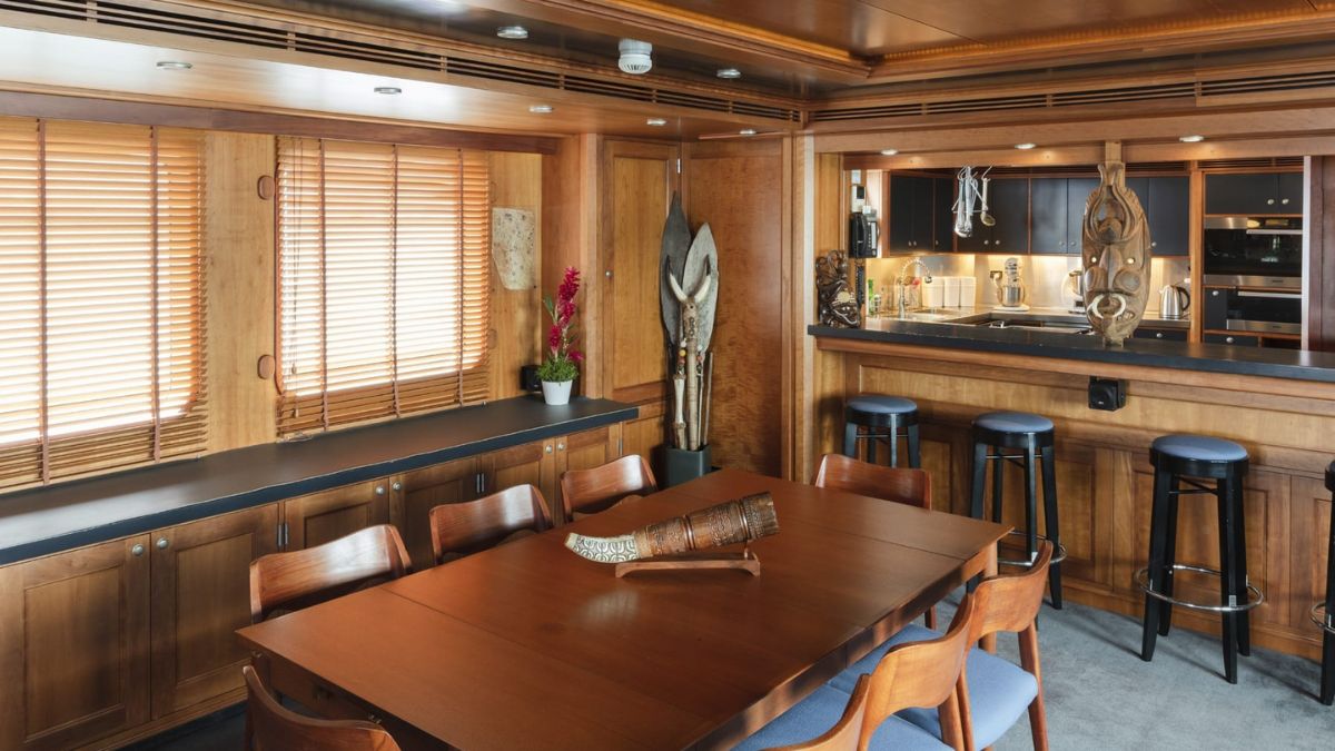 A modern wooden dining area with a large table surrounded by chairs. On one side, there are decorative objects and a small potted plant. The room features wooden blinds and a bar area with stools in the background.
