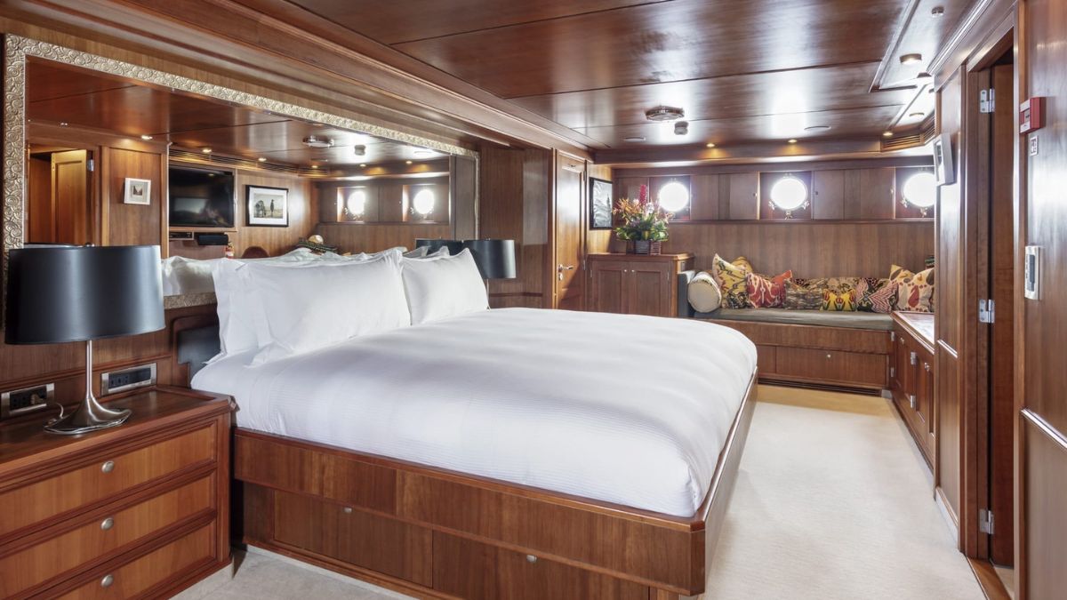 Luxurious yacht bedroom featuring a spacious king-size bed, warm wooden accents, and elegant decor. Round porthole windows let in natural light, complemented by stylish lamps and decorative pillows on a built-in seating area.
