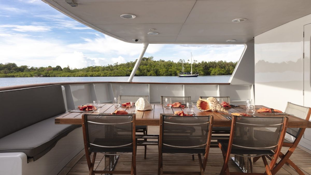 A dining table set with plates and glasses, surrounded by chairs, on a yacht's deck overlooking a calm water body and lush greenery.