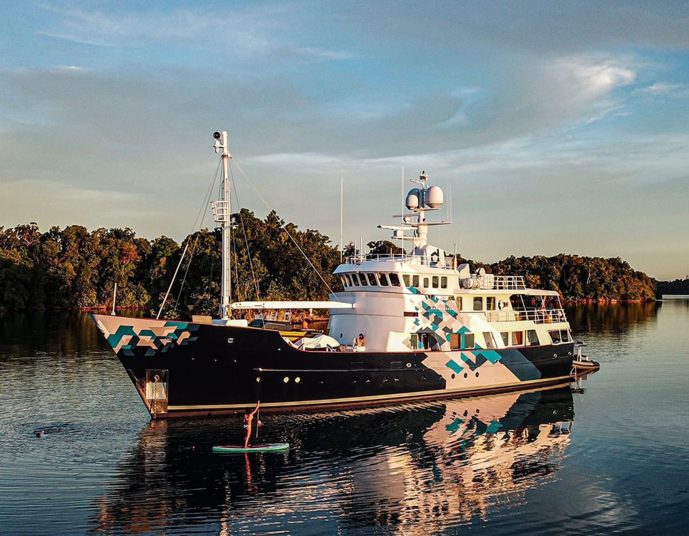 A large yacht with a striking blue and black camouflage pattern is moored in calm waters, surrounded by lush green trees. A person stands on a paddleboard nearby, enjoying the serene environment.
