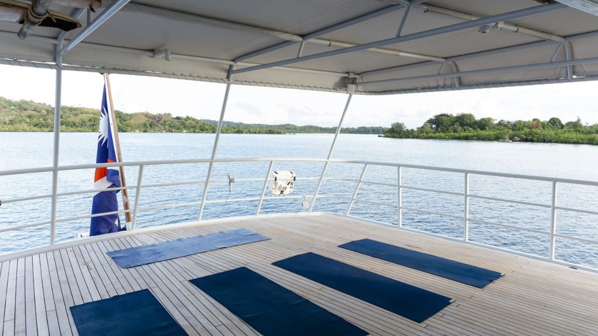A serene yoga deck on a boat, featuring several blue mats arranged on a wooden floor. In the background, calm waters and lush greenery are visible under a clear sky. A flag is visible on one side, gently waving in the breeze.
