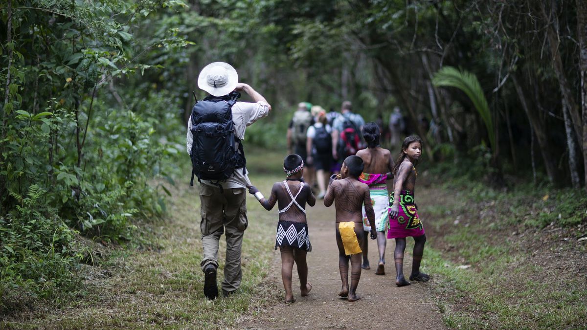 A group of people walking on a forest path, with an adult taking photos and children walking alongside, all surrounded by lush greenery.