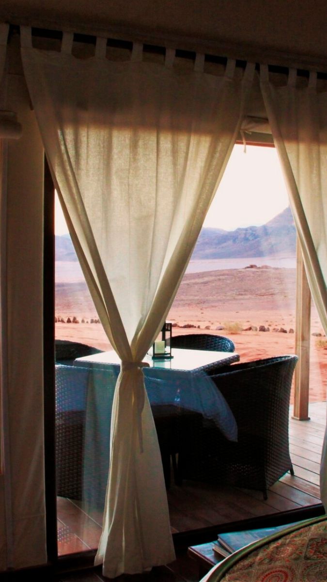 Sheer white curtains frame a doorway leading to a scenic outdoor view of a desert landscape with mountains in the background. A small table set for two is visible on the patio.