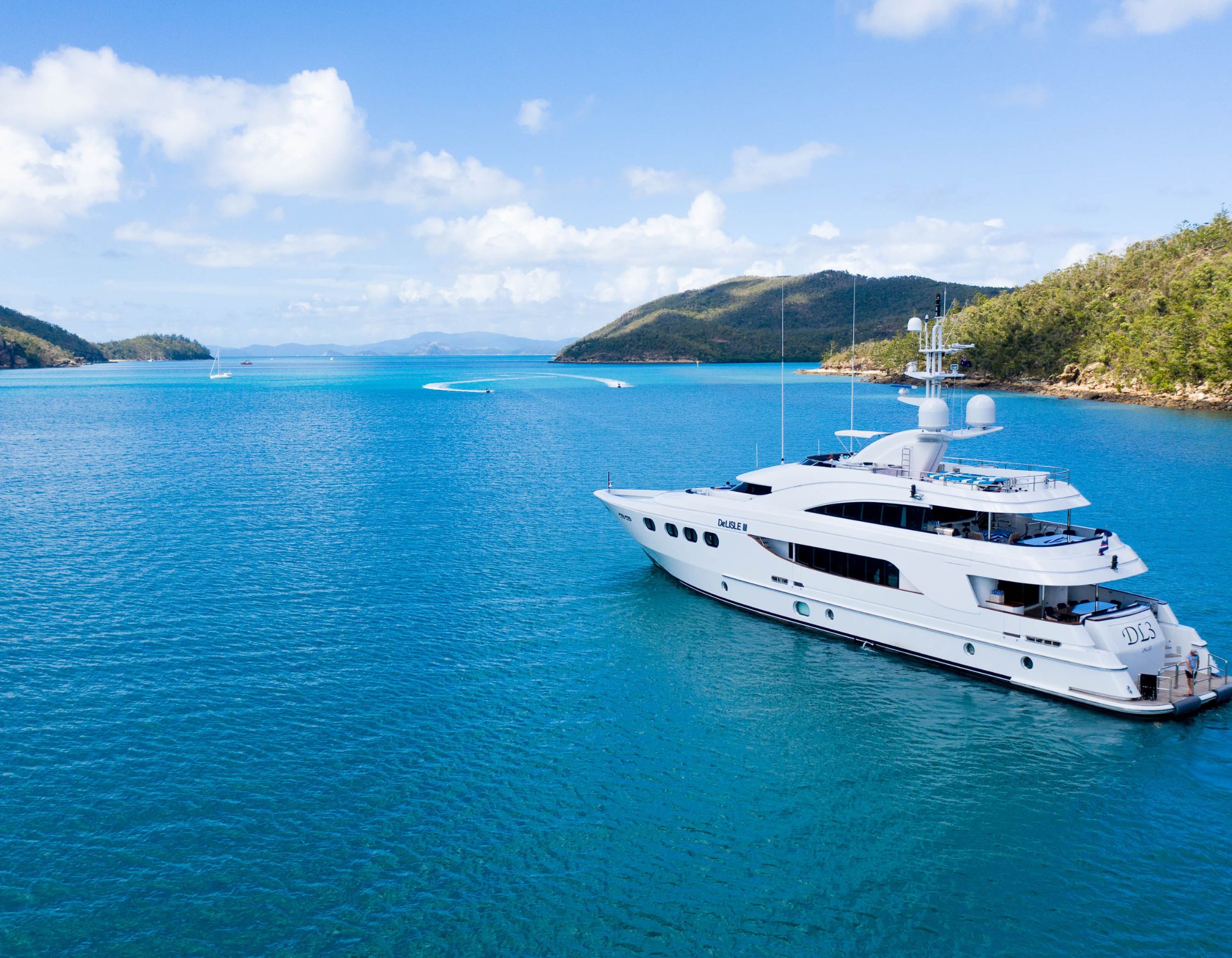 A luxurious white yacht anchored in turquoise waters, surrounded by lush green hills and clear blue skies.