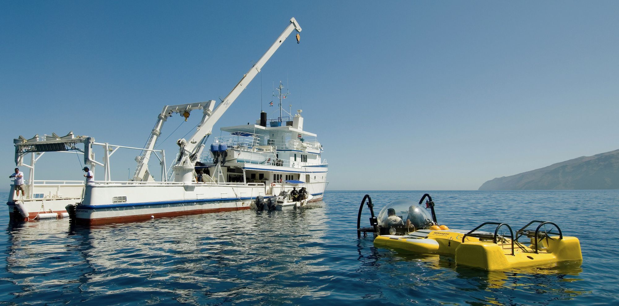 A large research ship is anchored in calm blue waters, with a crane extending over the side. Beside the ship, a yellow submersible is ready for exploration. A mountainous coastline is visible in the background under a clear blue sky.