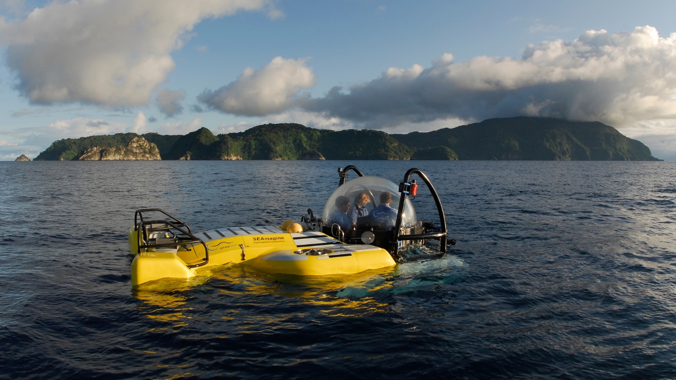 A yellow submersible is floating in calm waters, with two people inside observing the surroundings. Lush green islands rise in the background under a partly cloudy sky.