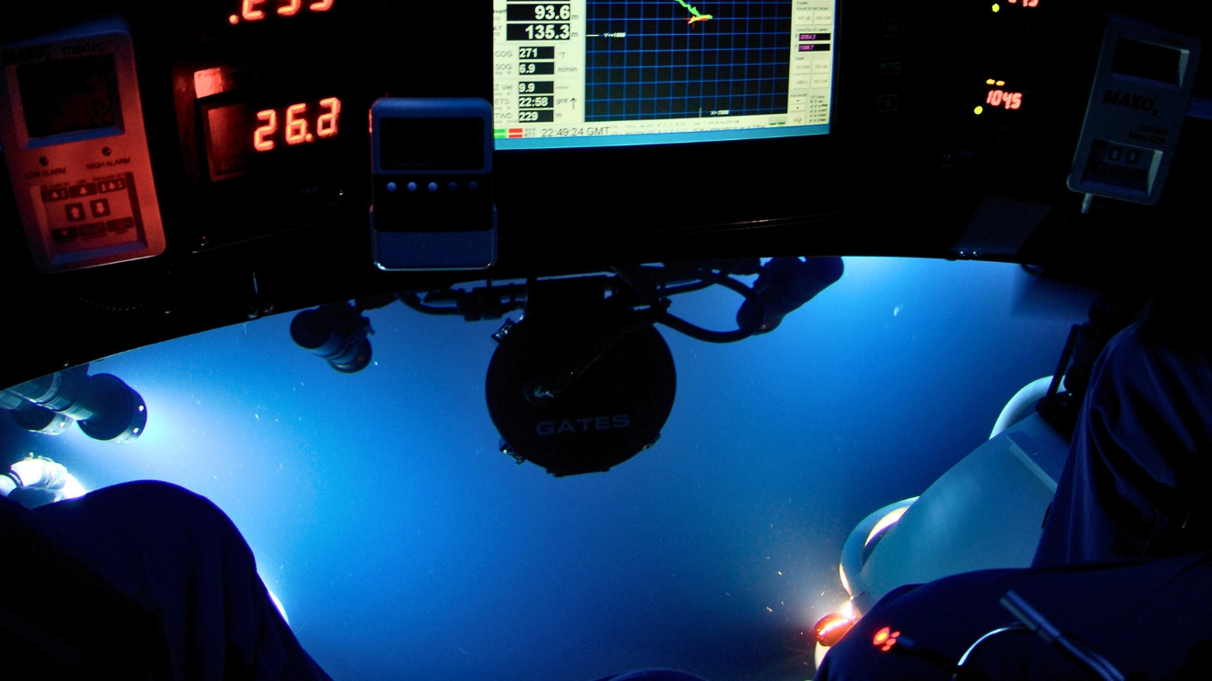 A control panel inside a submersible with various gauges and a digital display showing depth and coordinates. The view through the submersible's window reveals deep blue water with faint lights in the distance.