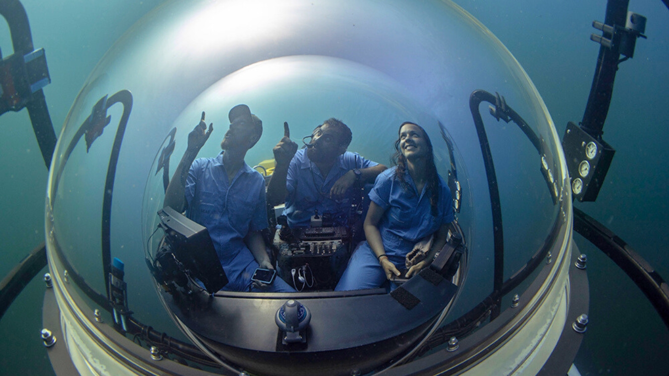Three people inside a submersible are exploring underwater. They are smiling and pointing at something outside the dome. The environment is blue and murky, suggesting depth in the ocean.