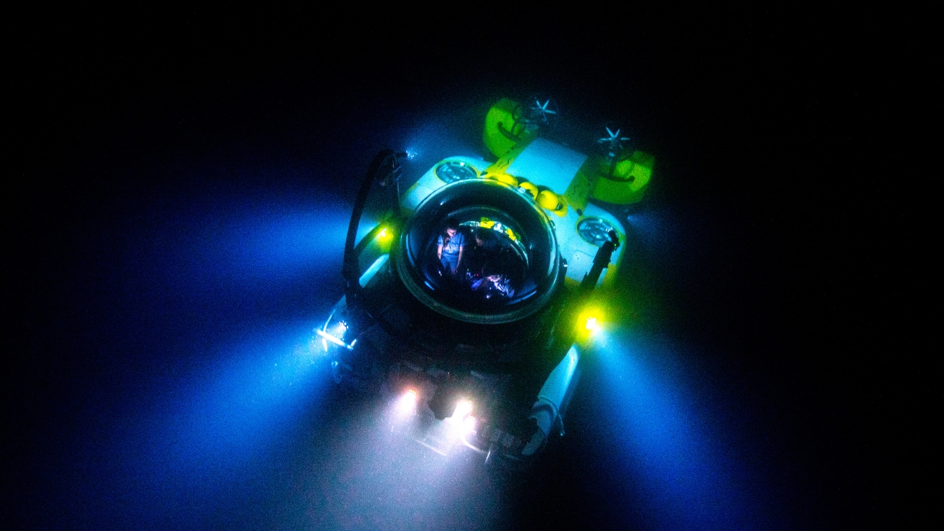 A submersible equipped with bright lights navigates through dark ocean waters, revealing its round cabin and propellers.