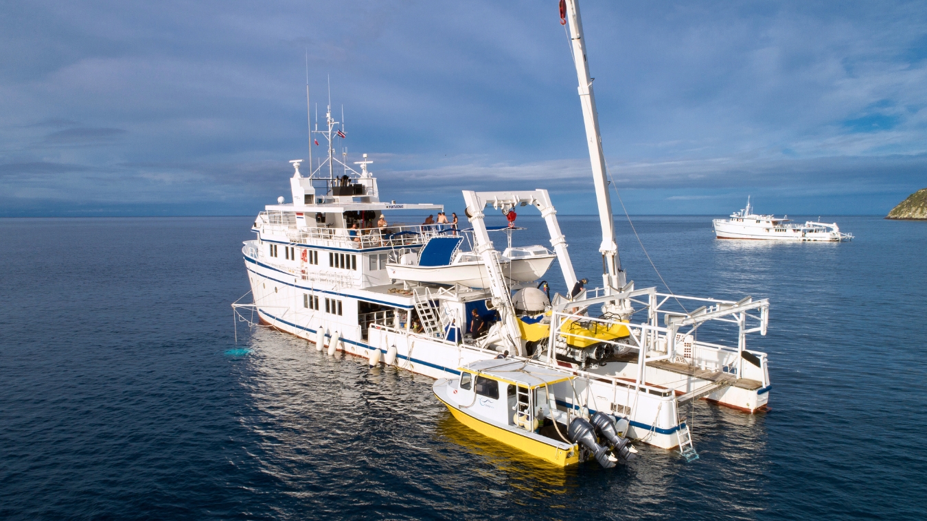 A large white research vessel is anchored in calm blue waters, with a smaller yellow boat docked alongside it. The sky is overcast with patches of blue. In the distance, another boat is visible on the water.