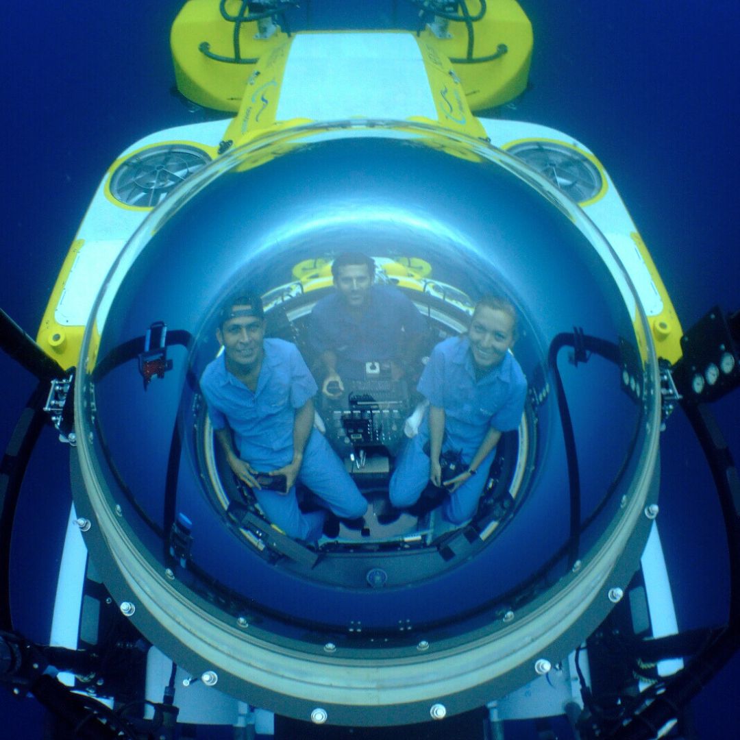 Three individuals seated inside a large submersible, with a round glass dome above that reflects their faces and surroundings. The interior is equipped with controls and displays, while deep blue water surrounds them.