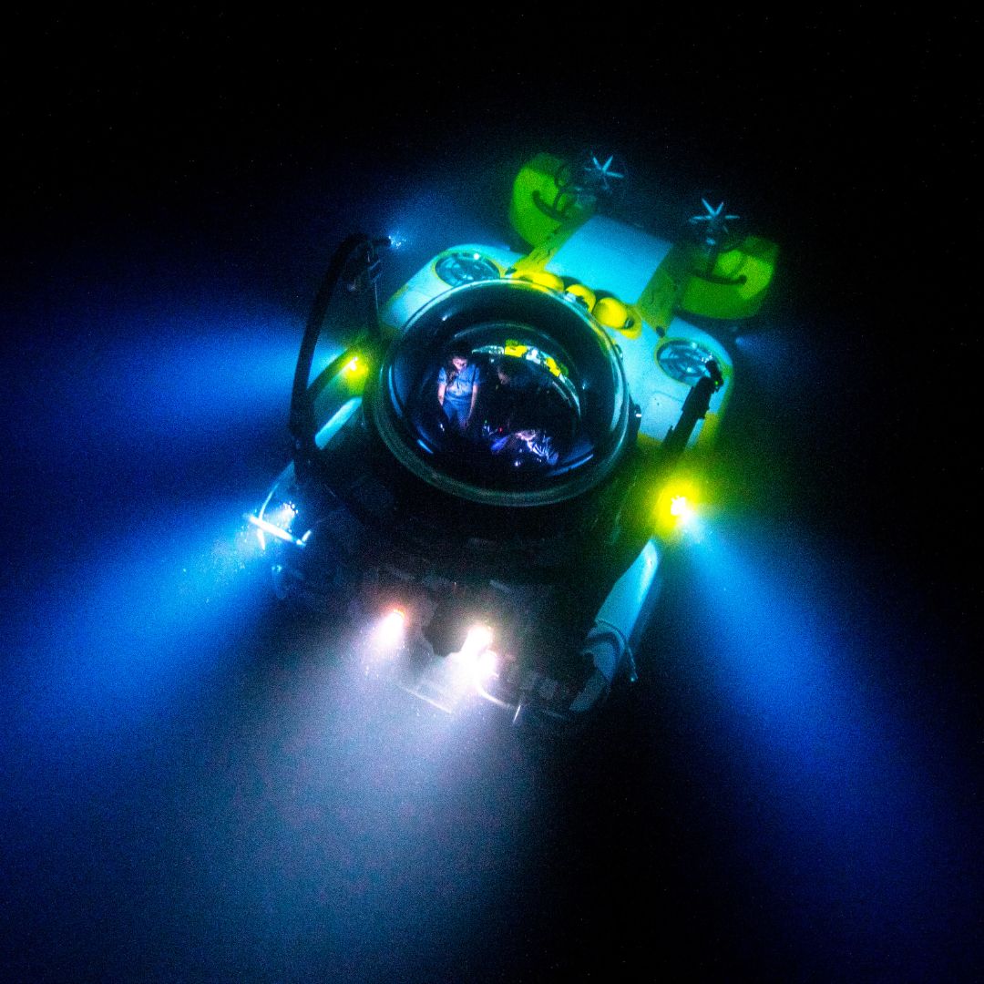 A submersible descends into dark water, illuminated by bright blue and yellow lights. The design features a large, dome-shaped viewing glass at the front, showcasing the underwater exploration.