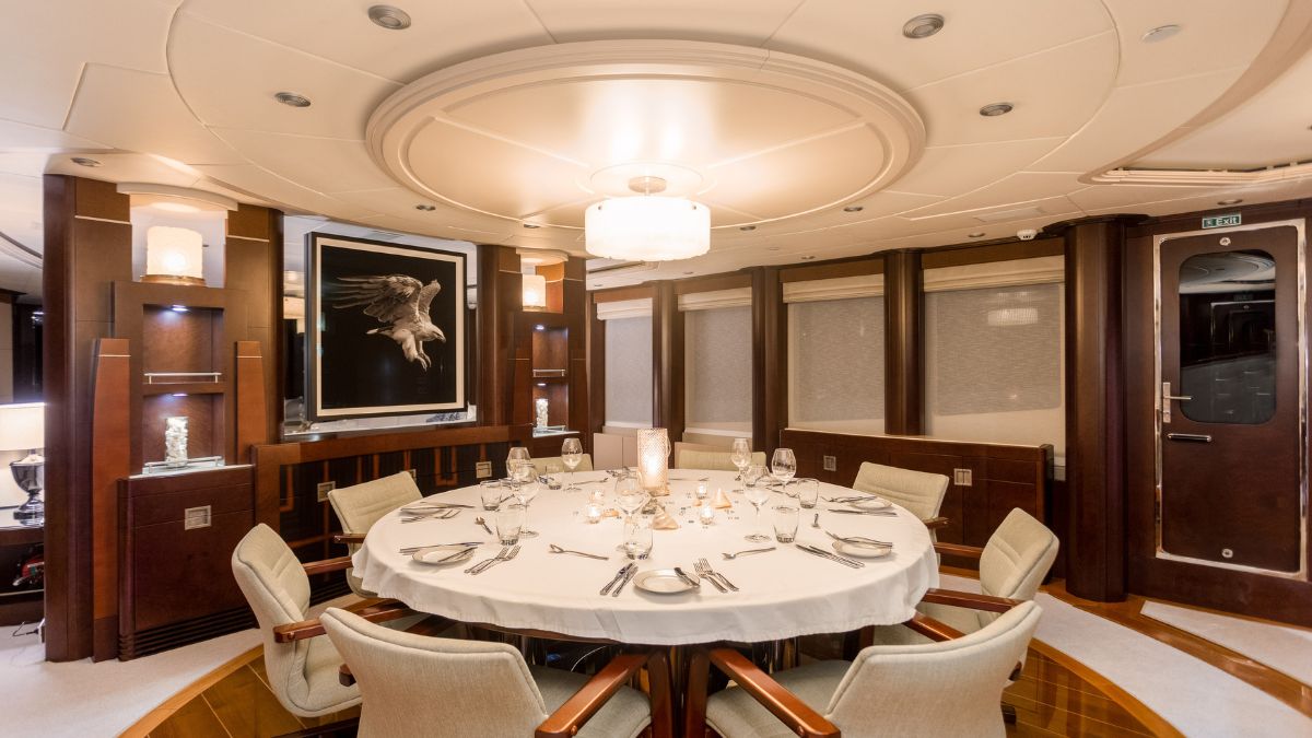 A luxurious dining room with a circular table set for a formal meal. The table is adorned with elegant place settings, and a large piece of art featuring an eagle hangs on the wall. Soft lighting creates a warm ambiance, complemented by rich wooden finishes.