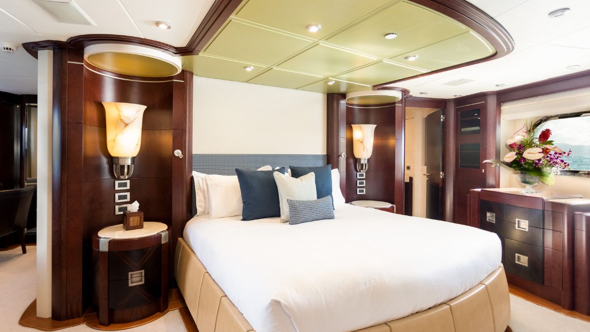 Luxurious yacht bedroom featuring a large bed with plush white bedding, accented by decorative pillows. Elegant wall sconces provide warm lighting, and the décor includes rich wooden finishes and a tasteful floral arrangement.