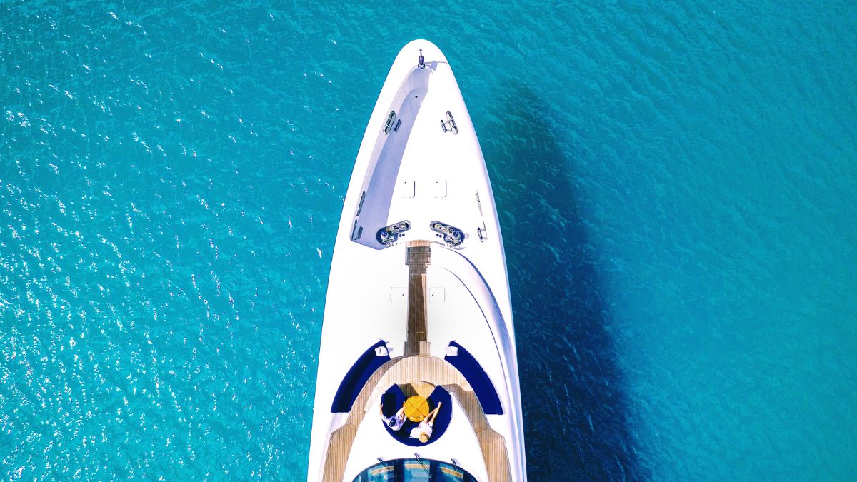 Aerial view of a luxurious yacht with a sleek white hull, sitting on vibrant turquoise water. A person relaxes on the deck, surrounded by a sunbathing area.
