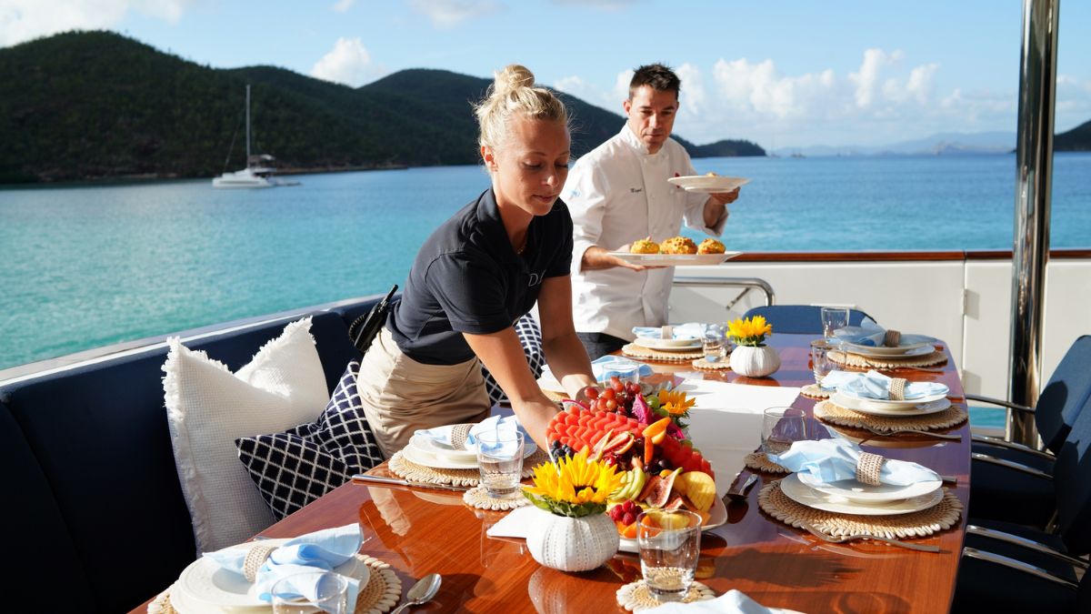 A woman sets a beautifully arranged dining table on a yacht, while a man carries a plate of food. The turquoise water and lush hills create a picturesque backdrop.