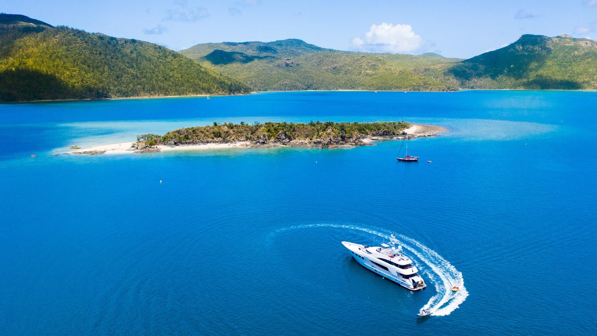 A luxury yacht glides through turquoise waters near a lush, green island surrounded by a sandy beach and calm waves. Majestic mountains rise in the background under a clear blue sky with a few clouds.