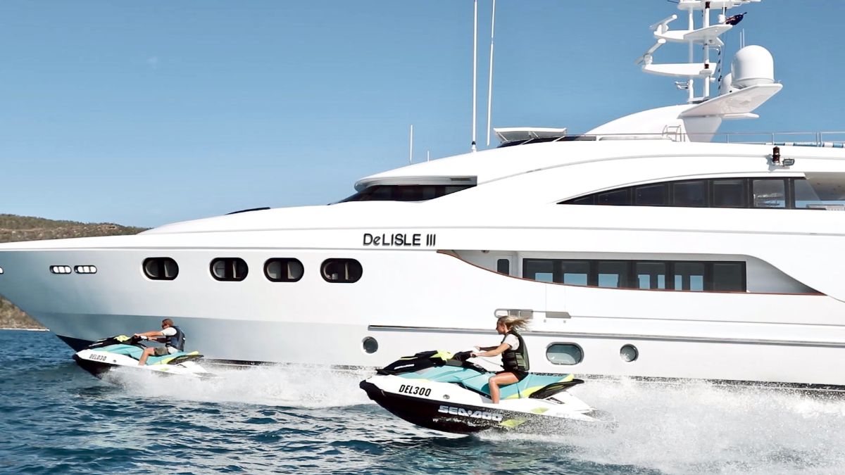 Two jet skis race beside a luxurious motor yacht named "DeLisle III," showcasing the vibrant blue water and clear skies.