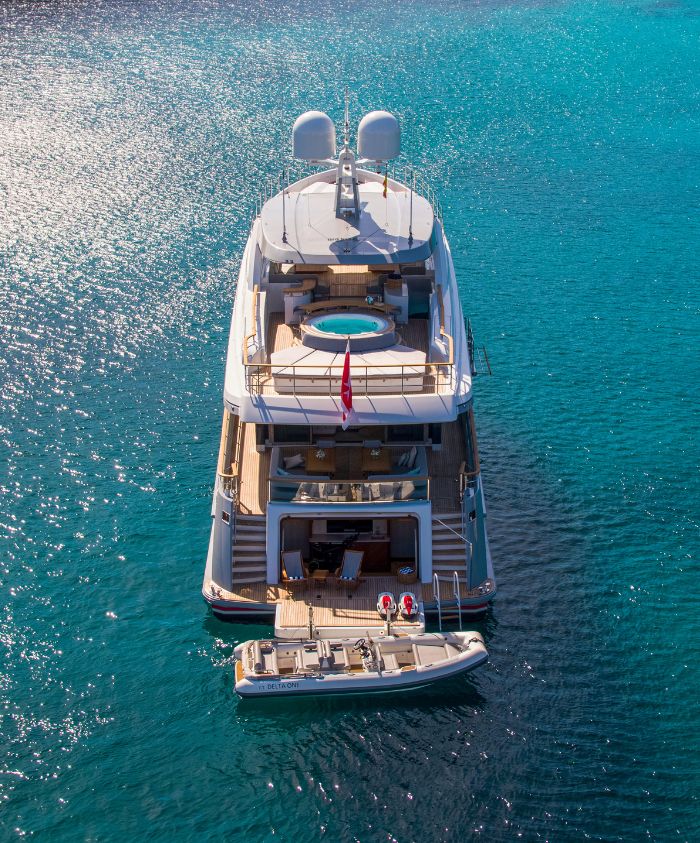 A luxurious yacht anchored in clear blue waters, featuring a rooftop pool and spacious decks. A smaller tender is moored alongside.