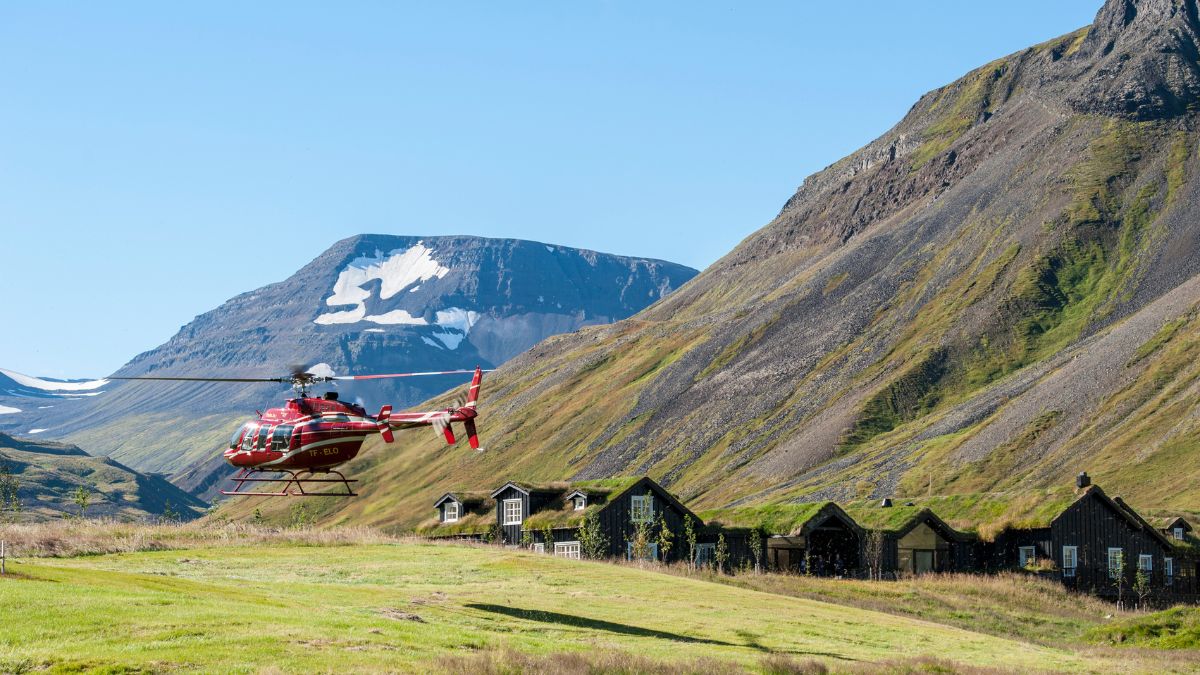 A red helicopter hovers near a cluster of rustic wooden cabins set against a backdrop of dramatic mountains and a bright blue sky. Snow caps some peaks in the distance.