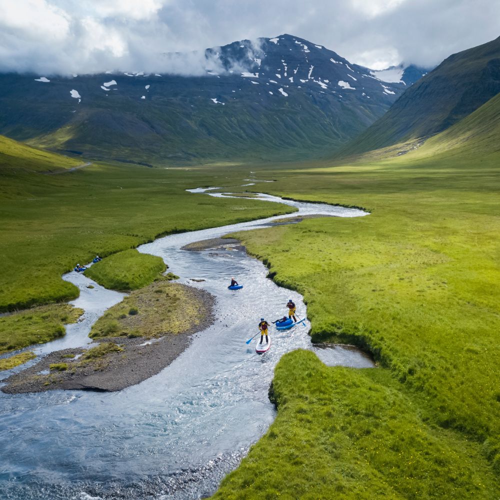Aerial view of a winding river flowing through lush green landscapes, with several individuals paddleboarding on the water. Majestic mountains are in the background under a partly cloudy sky.