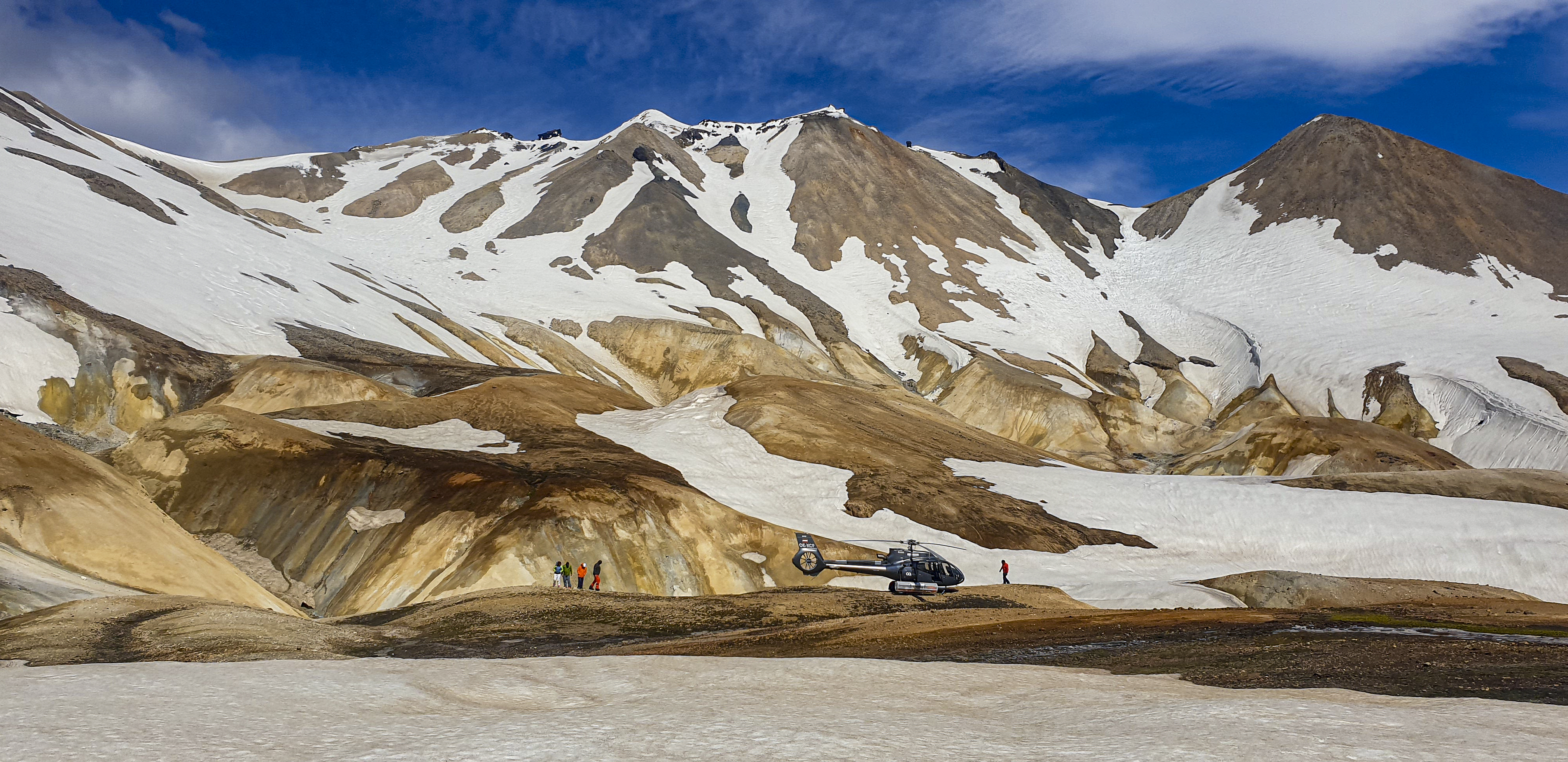 A helicopter is parked on a rocky, snow-covered landscape with mountains in the background. A few people are seen in the distance exploring the area. Clear blue skies and scattered clouds enhance the scenic view.
