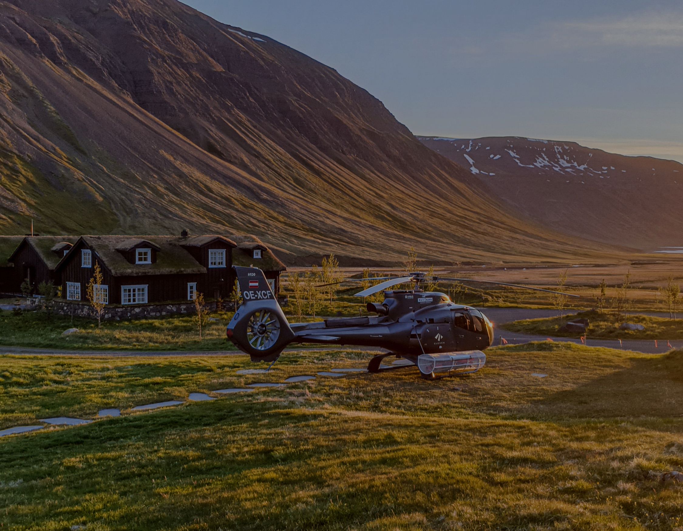 A helicopter is preparing for takeoff near a green grassy area with a rustic cabin in the background, set against a mountainous landscape. The scene is illuminated by warm, golden sunlight, suggesting early morning or late afternoon.