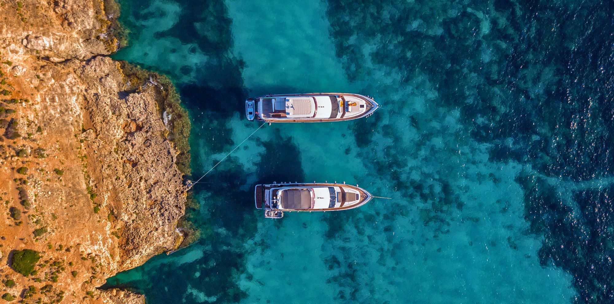 Two sleek yachts anchored near a rocky shoreline, surrounded by clear turquoise waters. The view is aerial, showcasing the vibrant contrast between the boats and the sea.